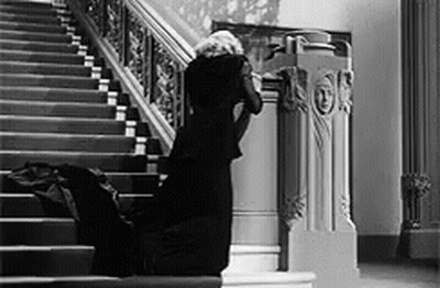 A Woman In A Black Dress Is Kneeling Down On The Stairs In A Black And White Photo . GIF