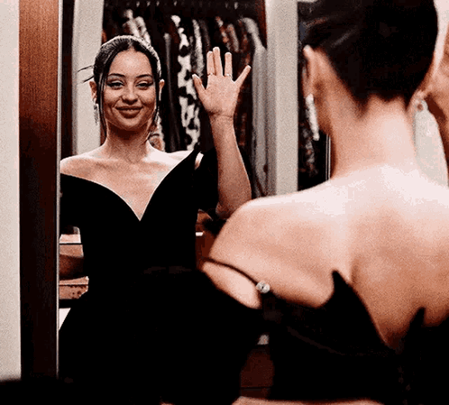 A Woman In A Black Dress Is Looking At Herself In The Mirror And Waving . GIF