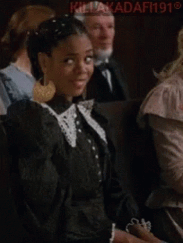 A Woman In A Black Dress Is Making A Funny Face While Sitting In A Crowd Of People . GIF