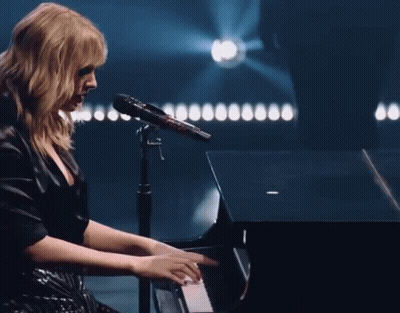 A Woman In A Black Dress Is Playing A Piano And Singing Into A Microphone GIF