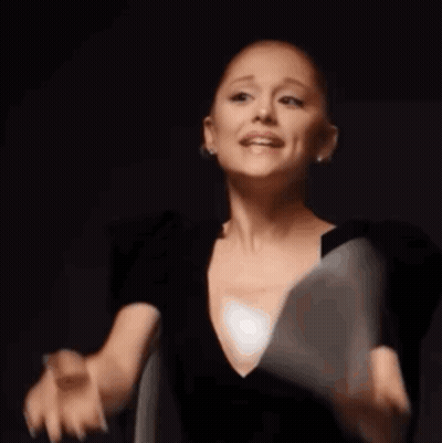 A Woman In A Black Dress Is Saying Gay While Holding A White Object In Her Hand GIF