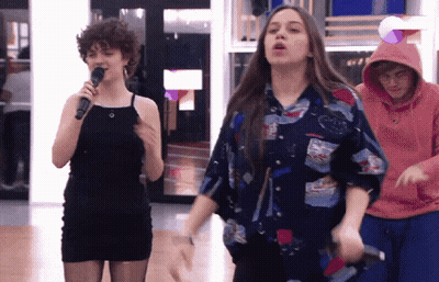A Woman In A Black Dress Is Singing Into A Microphone While A Man In A Pink Hoodie Watches GIF