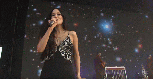 A Woman In A Black Dress Is Singing Into A Microphone While Standing In Front Of A Screen That Says Ella Talk GIF