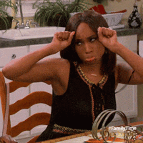 A Woman In A Black Dress Is Sitting At A Table With Her Hands On Her Head In A Kitchen .. GIF