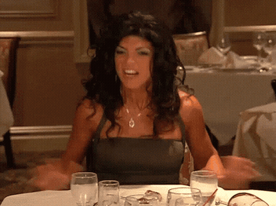 A Woman In A Black Dress Is Sitting At A Table With Wine Glasses GIF