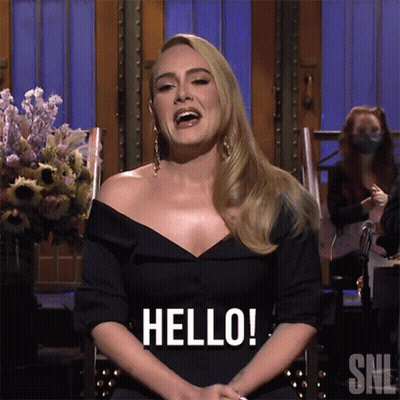 A Woman In A Black Dress Is Sitting In A Chair And Saying Hello GIF