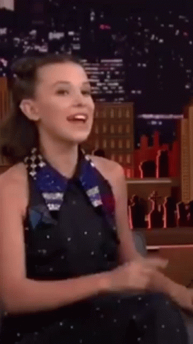 A Woman In A Black Dress Is Sitting In A Chair Laughing . GIF