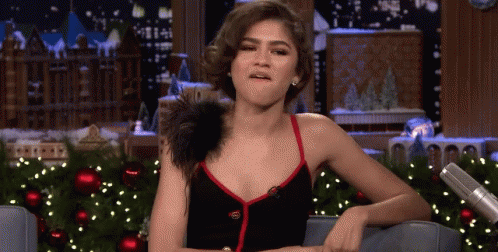 A Woman In A Black Dress Is Sitting In Front Of A Christmas Tree And Making A Funny Face . GIF