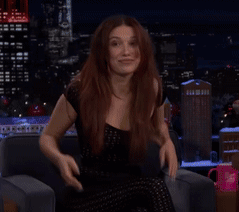 A Woman In A Black Dress Is Sitting On A Couch With Her Hands On Her Face GIF