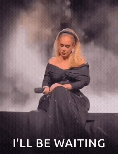 A Woman In A Black Dress Is Sitting On A Stage Holding A Microphone And The Words `` I 'Ll Be Waiting '' GIF