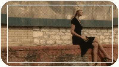 A Woman In A Black Dress Is Sitting On A Wall GIF
