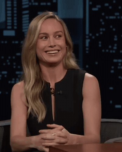 A Woman In A Black Dress Is Smiling With Her Hands Folded In Front Of Her GIF