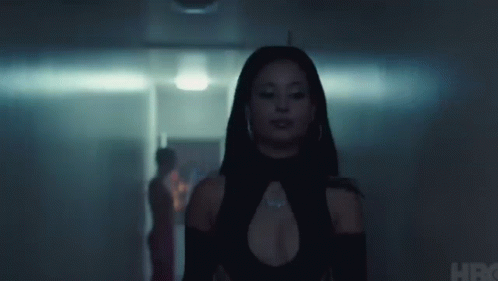 A Woman In A Black Dress Is Standing In A Dark Hallway Looking At The Camera . GIF