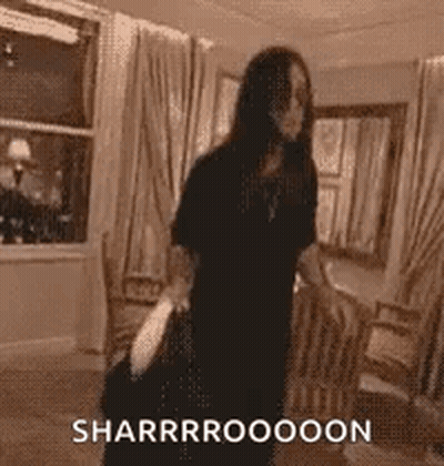A Woman In A Black Dress Is Standing In A Living Room Holding A Bag And A Purse . GIF