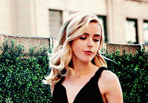 A Woman In A Black Dress Is Standing Next To A Hedge . GIF