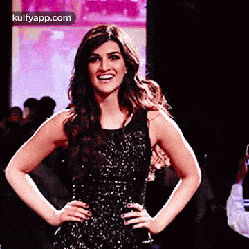 A Woman In A Black Dress Is Standing On A Runway With Her Hands On Her Hips And Smiling . GIF