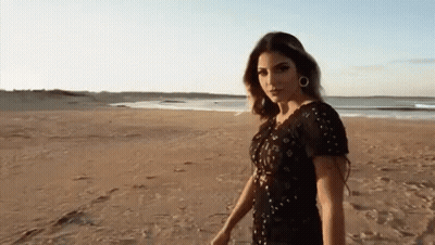 A Woman In A Black Dress Is Standing On A Sandy Beach GIF