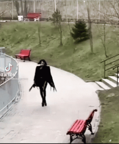 A Woman In A Black Dress Is Walking Down A Path With A Red Bench In The Background . GIF