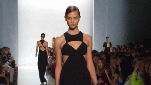 A Woman In A Black Dress Is Walking Down A Runway At A Fashion Show GIF