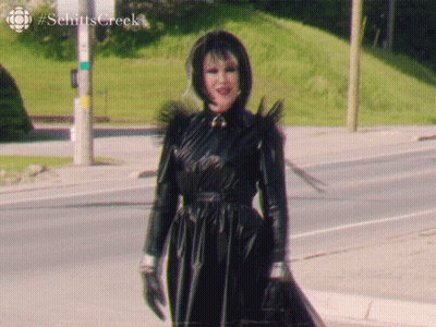 A Woman In A Black Dress Is Walking Down A Street With #Schitts Creek Written In The Corner GIF