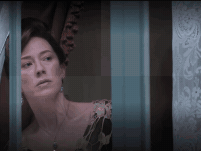 A Woman In A Black Dress Looks Out A Window With A Blue Curtain Behind Her GIF
