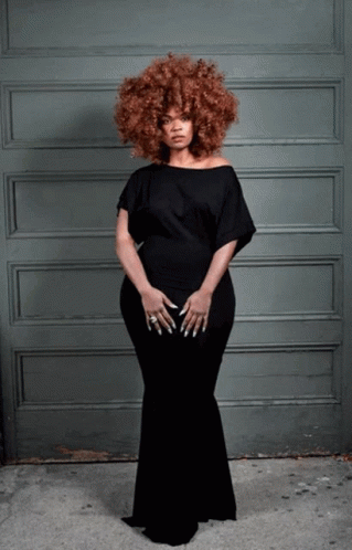 A Woman In A Black Dress Stands In Front Of A Gray Wall GIF