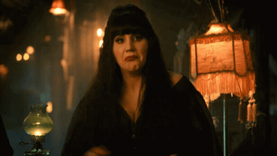 A Woman In A Black Dress Stands In Front Of A Lamp With A Fringed Shade That Says ' A ' GIF
