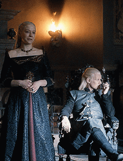 A Woman In A Black Dress Stands Next To A Man In A Chair Smoking A Cigarette GIF