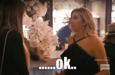 A Woman In A Black Dress Talks To Another Woman In Front Of A Sign That Says Ok On It GIF
