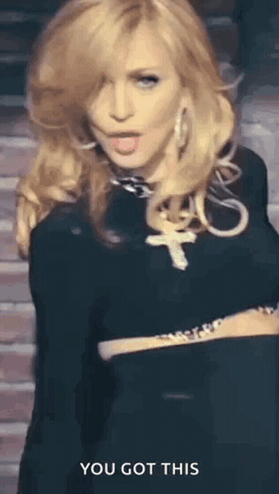 A Woman In A Black Dress With A Cross Necklace Is Standing In Front Of A Brick Wall And Says You Got This . GIF