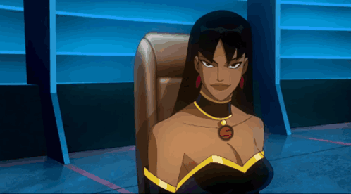 A Woman In A Black Dress With A Gold Necklace With The Letter S On It Sits In A Chair GIF