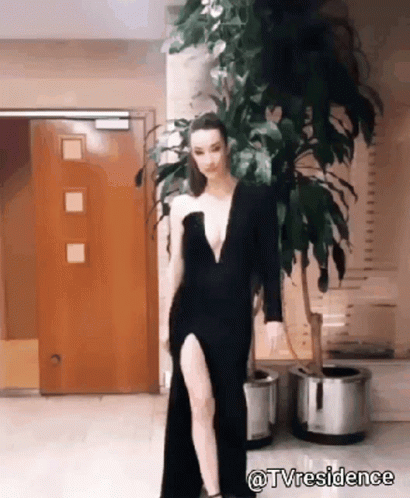 A Woman In A Black Dress With A High Slit Is Standing In Front Of A Plant . GIF