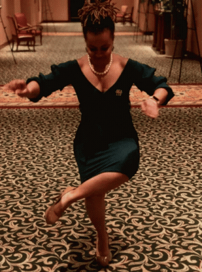 A Woman In A Black Dress With A Pearl Necklace And A Brooch With The Letter T On It Is Dancing On A Rug GIF