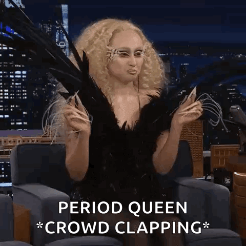 A Woman In A Black Dress With Feathers On Her Face Is Sitting In A Chair With The Words Period Queen Crowd Clapping . GIF