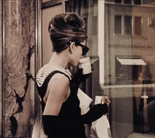 A Woman In A Black Dress With Pearls On The Back Is Drinking A Cup Of Coffee GIF