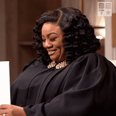 A Woman In A Black Gown Is Smiling In Front Of A Sign That Says Be Tm GIF