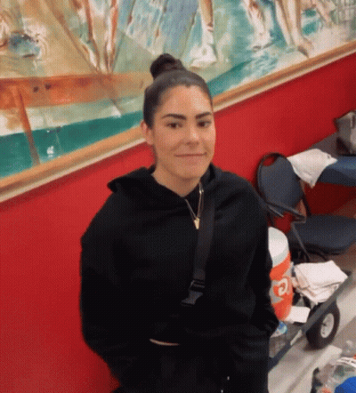 A Woman In A Black Hoodie Is Standing In Front Of A Red Wall And Smiling . GIF