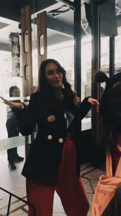 A Woman In A Black Jacket And Red Pants Is Standing In Front Of A Door GIF