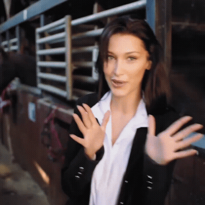 A Woman In A Black Jacket And White Shirt Is Waving Her Hands In Front Of A Horse Stable . GIF