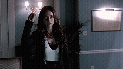 A Woman In A Black Jacket And White Top Stands In A Dark Room GIF