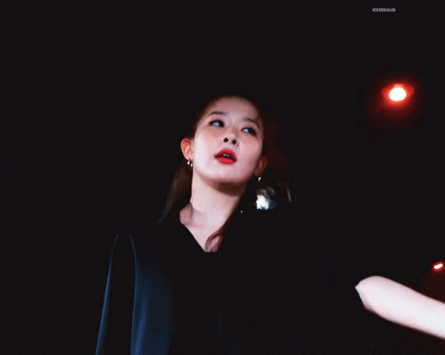 A Woman In A Black Jacket Is Dancing In A Dark Room With A Blue Light Behind Her GIF