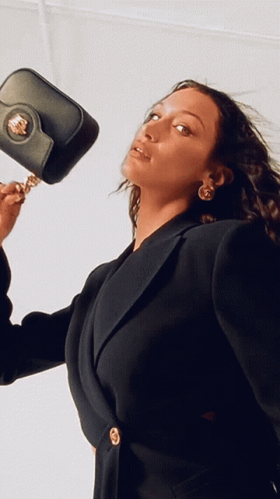 A Woman In A Black Jacket Is Holding A Black Purse On Her Shoulder GIF