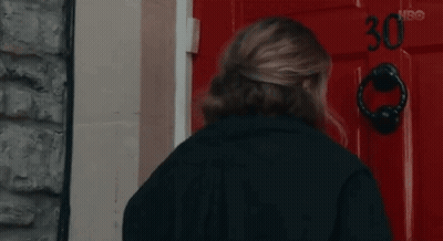 A Woman In A Black Jacket Is Knocking On A Red Door . GIF