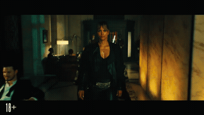 A Woman In A Black Jacket Is Standing In A Dark Room With A Sign That Says 18+ In The Corner GIF