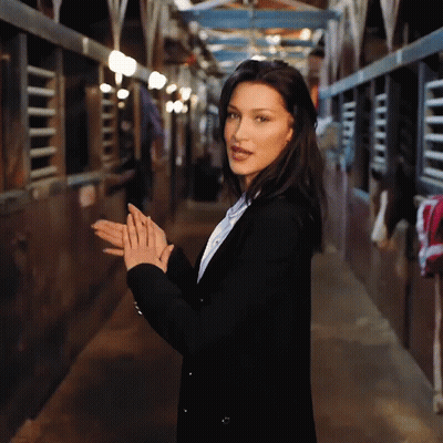 A Woman In A Black Jacket Is Standing In A Stable With Her Hands Folded GIF