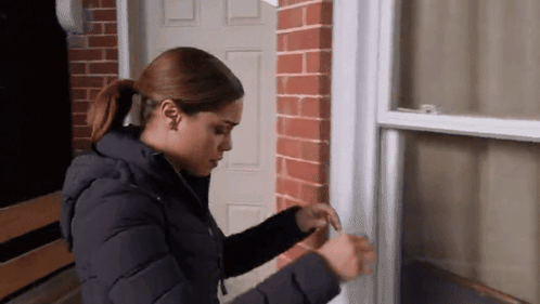 A Woman In A Black Jacket Is Standing In Front Of A Brick Door . GIF