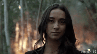 A Woman In A Black Jacket Is Standing In Front Of A Fire In The Woods . GIF