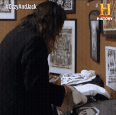 A Woman In A Black Jacket Is Standing In Front Of A Wall With Framed Pictures And The Words #Ozzyandjack Written On It GIF