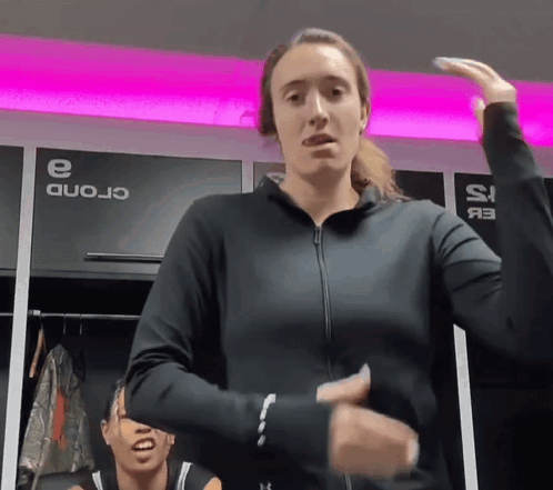 A Woman In A Black Jacket Stands In A Locker Room With A Sign That Says ' Crond ' On It GIF