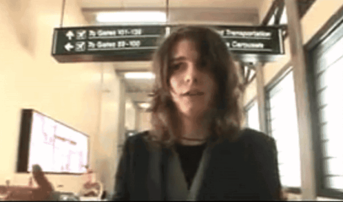 A Woman In A Black Jacket Stands In Front Of A Sign That Says Gates 41-130 GIF